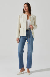 A woman is wearing a cream-colored, textured, long-sleeve, collarless jacket with gold buttons down the front and on the pockets. She has on a plain white top underneath. She is also wearing high-waisted, straight-leg blue jeans with a chain belt hanging from the waistband. Her shoes are pointed-toe, cream-colored high heels.